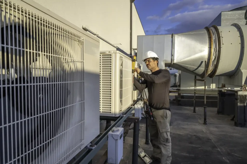 A male HVAC technician wearing a white hard hat, safety glasses, and casual work clothes is performing maintenance or repair on a bank of white commercial air conditioning units on a rooftop. He is using a handheld tool to work on the wiring or components of one of the units. Large metal ductwork and a clear sky with clouds are visible in the background, suggesting a commercial HVAC service setting.