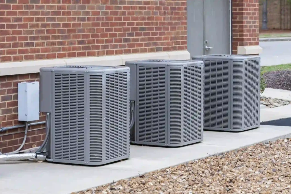 Three tall, grey, rectangular commercial air conditioning or heat pump condensing units are lined up on a concrete pad next to a red brick building. A white electrical disconnect box is mounted on the wall by the unit on the left. The ground in the foreground is covered with light brown gravel, indicating an external HVAC installation area.