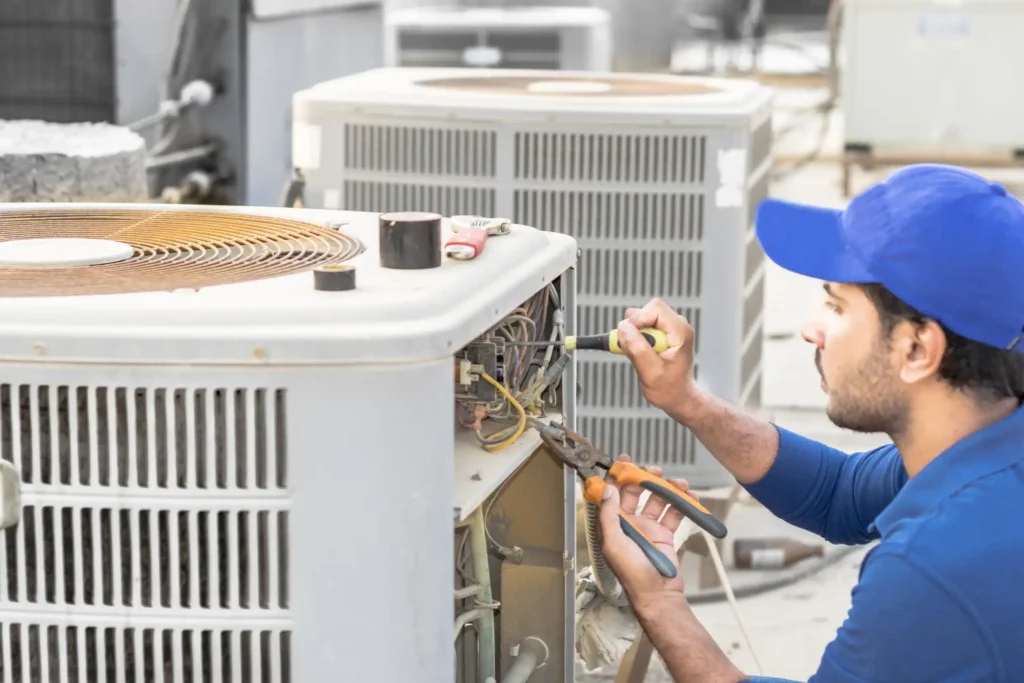 A male HVAC technician wearing a blue shirt and a blue baseball cap is servicing an open commercial air conditioning unit on a rooftop. He is using a screwdriver in one hand and a pair of pliers in the other to work on the exposed wiring and electrical components inside the unit. The top of the unit has a visible fan, and a second, larger HVAC unit is blurred in the background.