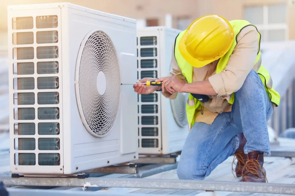A male HVAC technician wearing a bright yellow hard hat and a high-visibility safety vest is kneeling down to work on a white commercial air conditioning unit on a rooftop. He is using a screwdriver to remove or tighten a screw near the circular fan grille. The bright sunlight suggests an outdoor maintenance or repair job, with another unit visible behind him.