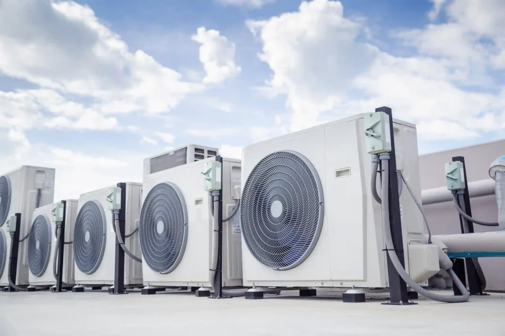 A low-angle shot of a row of white commercial air conditioning condensing units installed on a rooftop against a bright blue sky with white clouds. Each unit features a large circular fan grille and an adjacent black stand holding an electrical disconnect box. This image clearly depicts a commercial HVAC system installation.