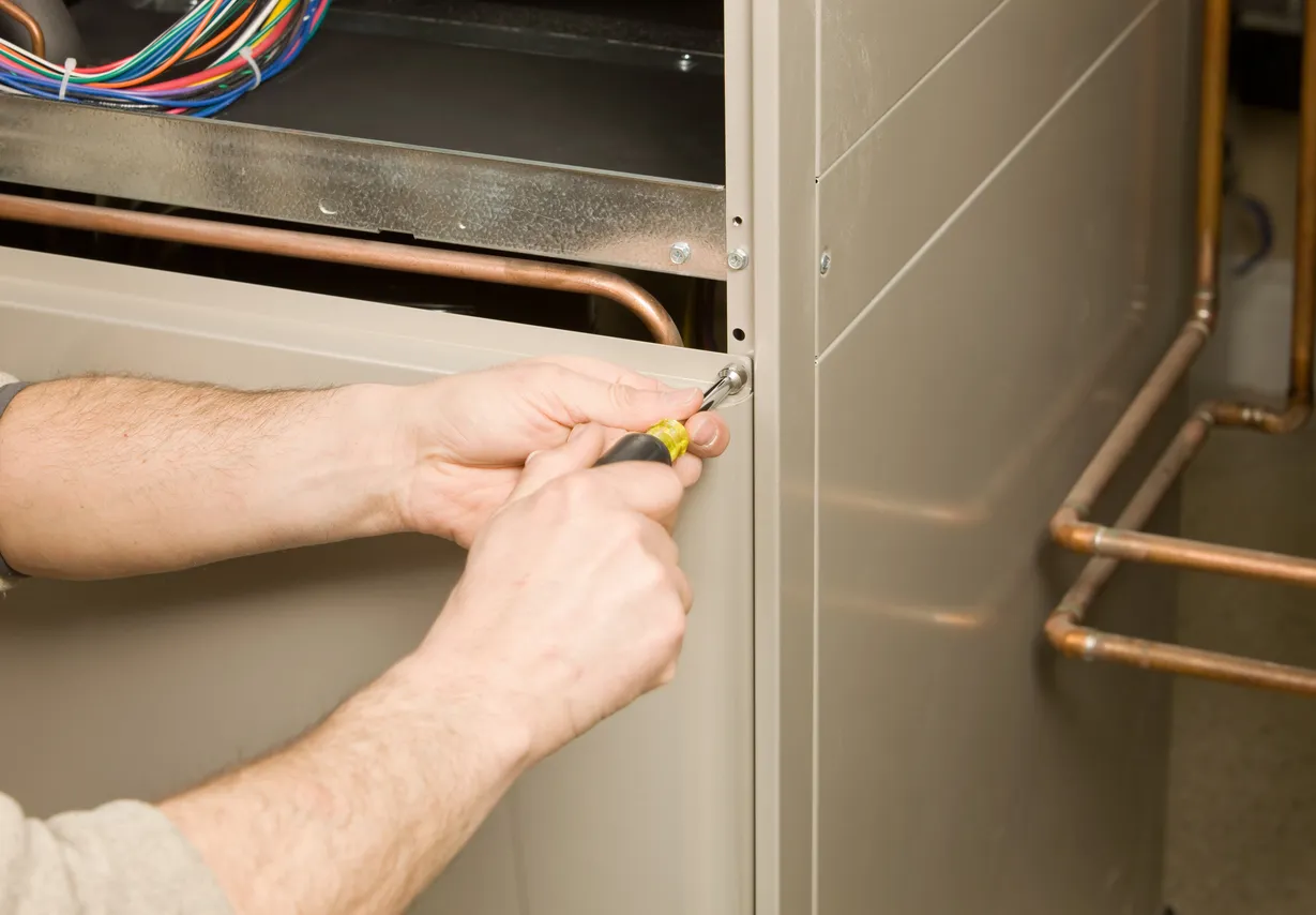 A close-up shot of a person using a screwdriver to remove a screw from the metal casing of a furnace, part of a home repair or maintenance task.